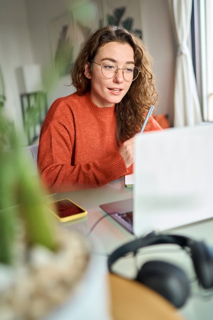 Woman with orange sweater and curly hair and glasses talking to laptop