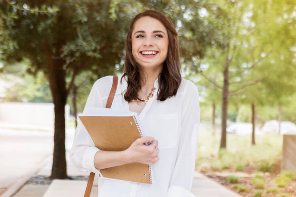 Woman in a park in white shirt holding a notebook and smiling