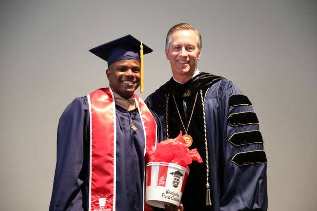 Kevon Pascoe stands with WGU President Scott Pulsipher. He is wearing graduation gowns and a red sash and holding a KFC bucket with his picture on it.