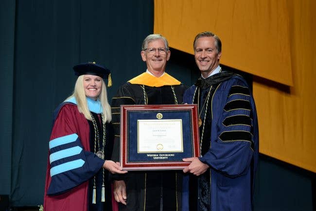 Left to right: Provost Courtney Hills McBeth, Dr. Charles Sorenson, and President Scott Pulsipher wearing commencement regalia and standing on a stage, with Sorenson holding a framed copy of his honorary degree.