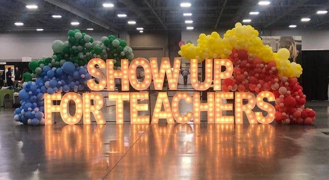 Light bulb sign that spells out Show Up for Teachers surrounded by multicolored balloons.